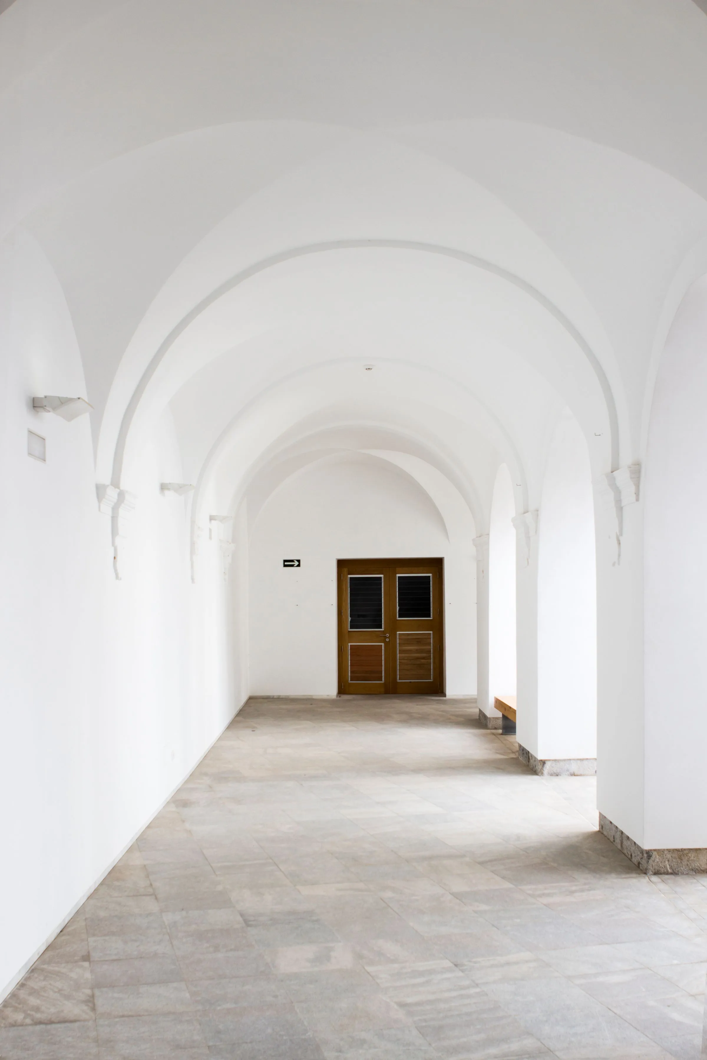 Pasillo blanco con arcos y una puerta de madera al fondo, con luz natural.